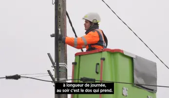 un ouvrier travail sur un poteau électrique, dans une cabine élévatrice, il porte un casque, des gants, et sa tenue de protection