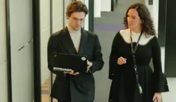 deux salariés, un homme et une femme, marchent et discutent dans un couloir d'entreprise, l'homme porte un ordinateur portable tout en marchant